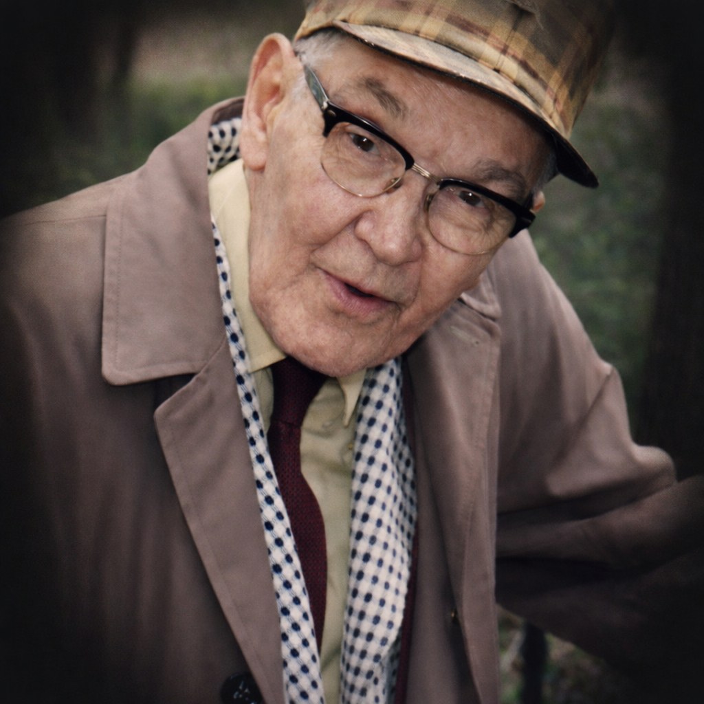 Portrait of Carroll Gunn in later life, wearing a plaid cap, glasses, a tan overcoat and a patterned scarf