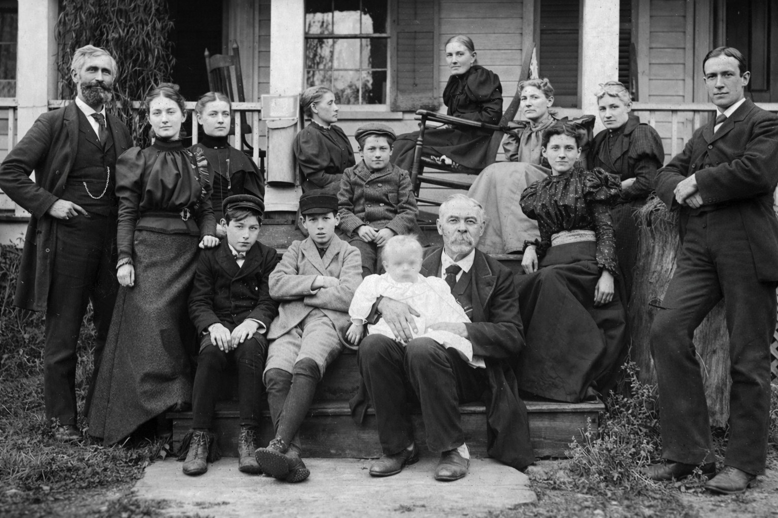 Gunn family group of about fifteen people posed on the steps and porch of a frame house; in the foreground an older bearded man holds an infant in a white gown